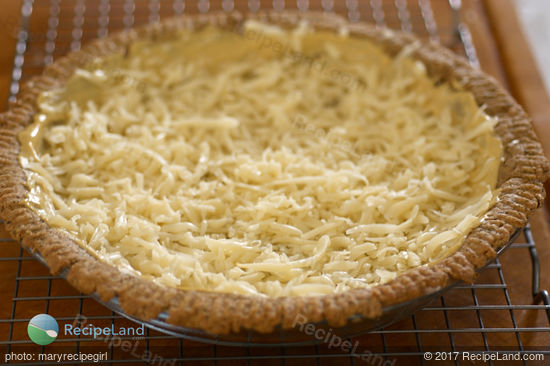 Blind baked herbed pie crust spread with Dijon mustard and partially filled with shredded mozzarella cheese in a glass pie plate on a wire rack.