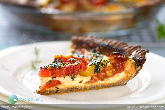 A juicy slice of Cheesy Herbed Fresh Heirloom Tomato Pie on a white plate with whole pie in the background and serving fork off to the side.