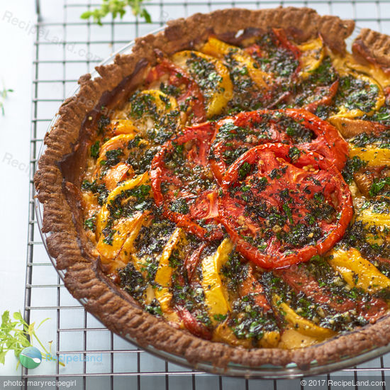 Overhead of a whole tomato pie.  Cheesy Herbed Heirloom Tomato pie topped with garlic herb oil in a glass pie plate on a wire rack on a blue background with fresh herbs scattered about.