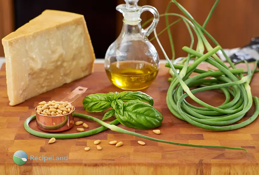 Garlic scape pesto ingredients, garlic scapes, basil, toasted pinenuts, olive oil and parmesan cheese arranged on a wood cutting board.