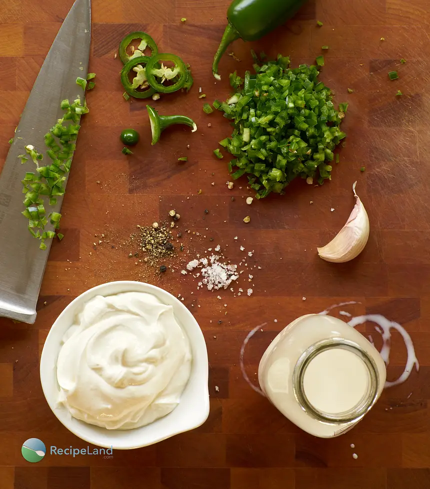 Ingredients to prepare Jalapeño cream sauce on a wood cutting board with a chef’s knife. Jalapeño peppers, salt, cracked black pepper, cream, sour cream, garlic.